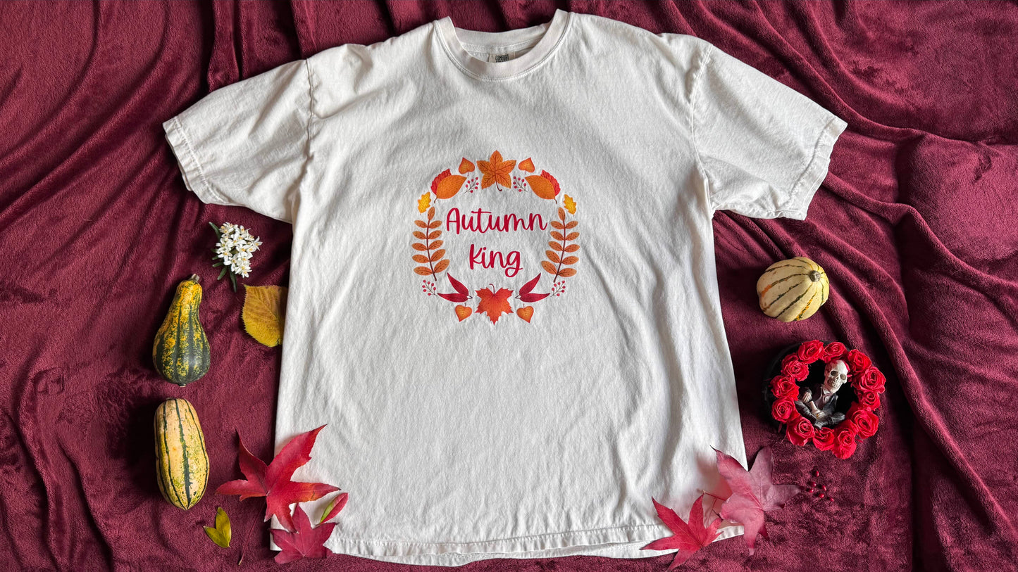 White t-shirt with 'Autumn King' text surrounded by cartoon autumn leaf's on a maroon fabric background with decorative elements.